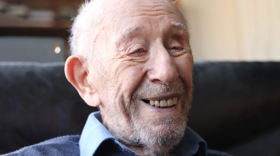 Eric Sanders, face of an old, grey haired, man, he has a wide smile on his face; he is wearing a blue collared shirt and a dark blue sweater